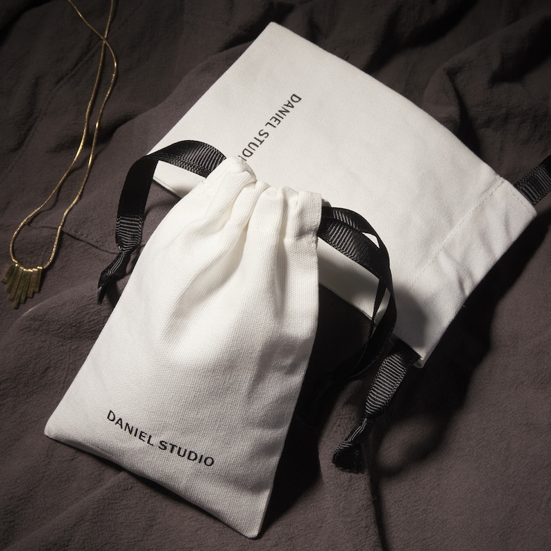 May include: A white drawstring pouch and a matching flat bag with black ribbon ties, both bearing the text "DANIEL STUDIO" in black. A delicate gold necklace with a geometric pendant lies nearby. The items rest on a dark gray fabric surface.