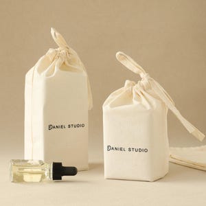 May include: Two off-white fabric drawstring pouches with the text DANIEL STUDIO printed in black on the front. A small clear glass bottle with a black dropper cap is positioned horizontally in front of the taller pouch on the left side.