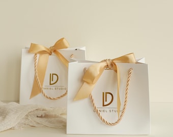 May include: Two white square gift bags featuring gold rope handles and a gold satin ribbon bow tied at the top. Each bag displays a gold logo with the letters LD and the text DANIEL STUDIO printed in a clean, centered font on the front surface.