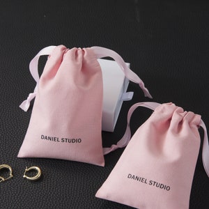 May include: Two light pink drawstring pouches with white ribbon ties, each printed with "DANIEL STUDIO" in black. A white box with a ribbon is visible behind the pouches. Gold hoop earrings are in the lower left corner.