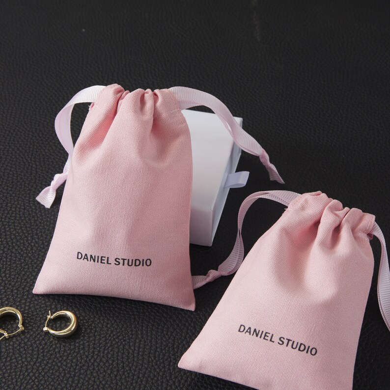 May include: Two light pink drawstring pouches with white ribbon ties, each printed with "DANIEL STUDIO" in black. A white box with a ribbon is visible behind the pouches. Gold hoop earrings are in the lower left corner.