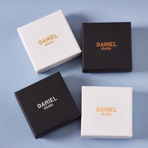 May include: Four square boxes, two white and two black, with the text "DANIEL studio" in gold lettering on each box.
