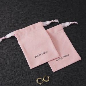 May include: Two light pink drawstring pouches with the text "DANIEL STUDIO" printed in black. The pouches have white ribbon drawstrings. A pair of gold hoop earrings rests near the pouches on a black textured surface.