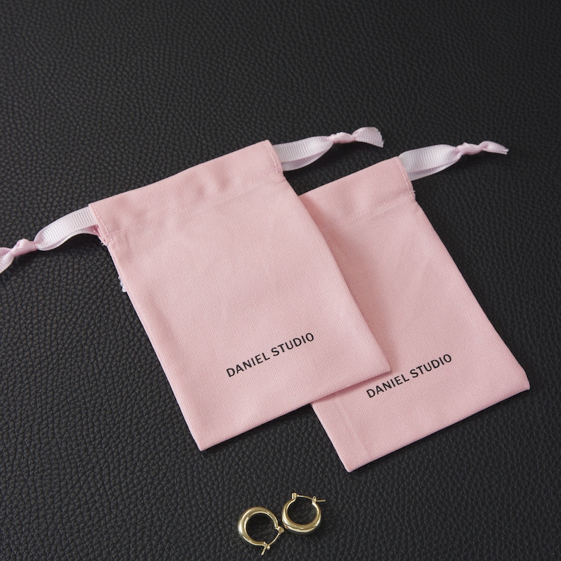May include: Two light pink drawstring pouches with the text "DANIEL STUDIO" printed in black. The pouches have white ribbon drawstrings. A pair of gold hoop earrings rests near the pouches on a black textured surface.