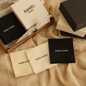 May include: A close-up of three white and black fabric pouches with the text "DANIEL STUDIO" printed on them. The pouches are on a beige fabric surface.