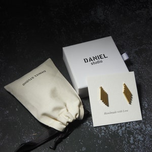 May include: Gold-colored geometric earrings on a white card with the text "Handmade with Love." The earrings are displayed with a white box and a beige drawstring bag, both labeled "DANIEL studio."