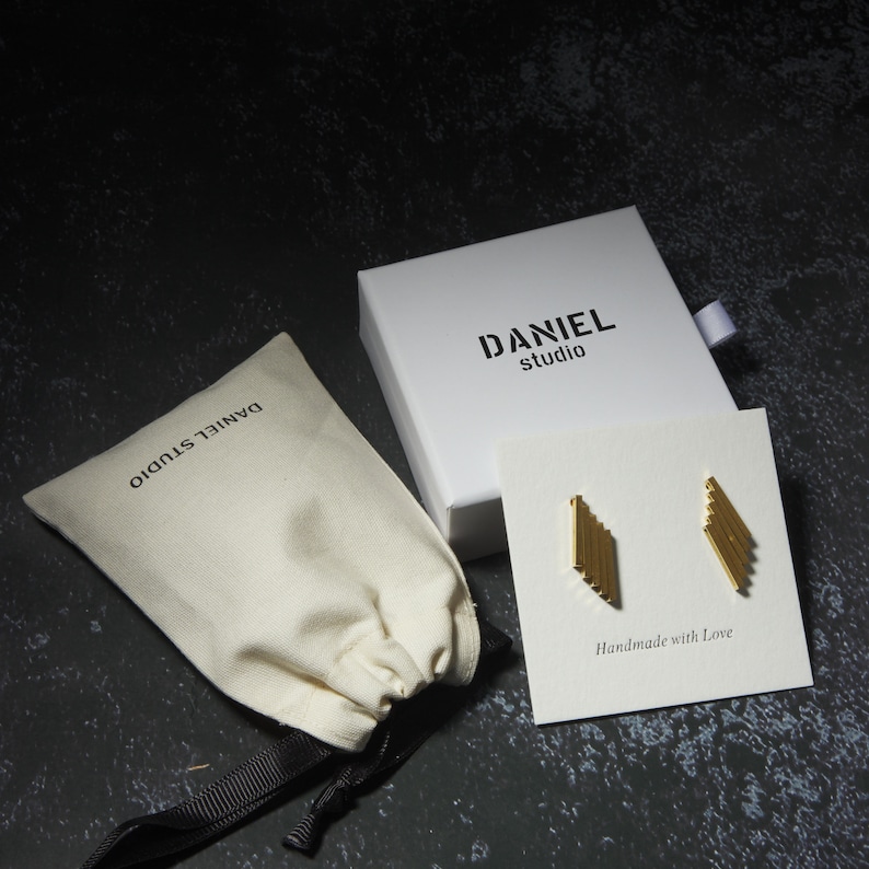 May include: Gold-colored geometric earrings on a white card with the text "Handmade with Love." The earrings are displayed with a white box and a beige drawstring bag, both labeled "DANIEL studio."