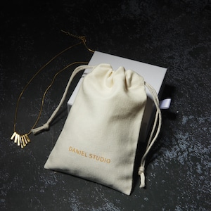 May include: A gold-colored necklace with a fan-shaped pendant rests beside a cream-colored drawstring bag and a white box. The bag is printed with "DANIEL STUDIO" in gold lettering. The background is a dark, textured surface.