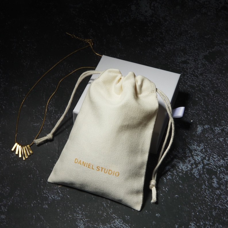 May include: A gold-colored necklace with a fan-shaped pendant rests beside a cream-colored drawstring bag and a white box. The bag is printed with "DANIEL STUDIO" in gold lettering. The background is a dark, textured surface.