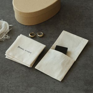 May include: A pair of gold hoop earrings, a small black rectangular box, and two off-white fabric pouches. One pouch features the black printed text DANIEL STUDIO. An oval-shaped tan cardboard box is positioned in the upper portion of the frame.