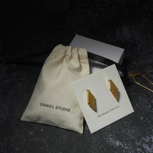 May include: Gold-colored earrings with a linear design are displayed on a white card that reads "Handmade with Love." The earrings are presented with a drawstring bag that says "DANIEL STUDIO" and a white box with a black ribbon.