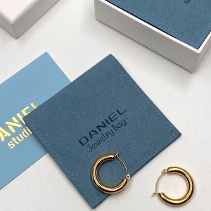 Może przedstawiać: Złote kolczyki koła na niebieskim aksamitnym woreczku na biżuterię z napisem "DANIEL Jewelry Bag".