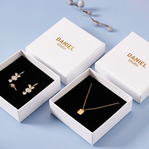 May include: Three white gift boxes with black velvet interiors. The boxes are labeled "DANIEL studio". The boxes contain jewelry: a pair of earrings, a ring, and a necklace.