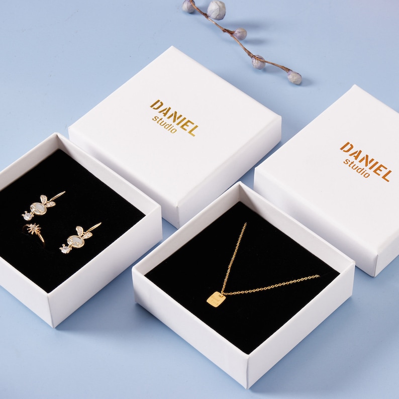 May include: Three white gift boxes with black velvet interiors. The boxes are labeled "DANIEL studio". The boxes contain jewelry: a pair of earrings, a ring, and a necklace.