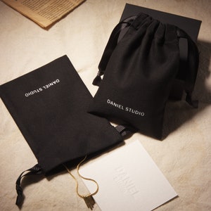 May include: An overhead shot showcases a gold necklace with a tassel pendant, displayed alongside black drawstring pouches and a white card, all bearing the "DANIEL STUDIO" logo. The items are arranged on a neutral-toned surface.
