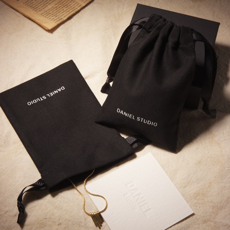 May include: An overhead shot showcases a gold necklace with a tassel pendant, displayed alongside black drawstring pouches and a white card, all bearing the "DANIEL STUDIO" logo. The items are arranged on a neutral-toned surface.