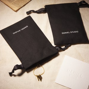 May include: Two black drawstring pouches with the text "DANIEL STUDIO" in white. A gold necklace with rectangular pendants is partially visible. A white card with the embossed text "DANIEL studio" is also present.