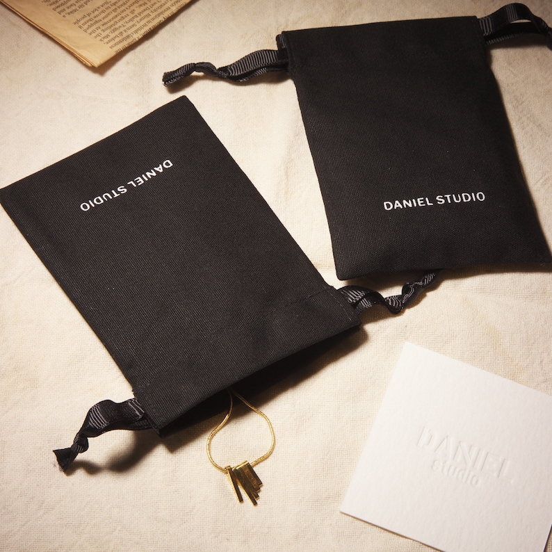 May include: Two black drawstring pouches with the text "DANIEL STUDIO" in white. A gold necklace with rectangular pendants is partially visible. A white card with the embossed text "DANIEL studio" is also present.