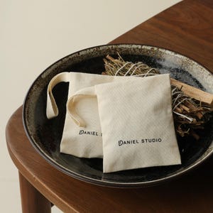 May include: Two off-white canvas drawstring pouches featuring the black text DANIEL STUDIO printed on the front. The pouches are placed inside a dark, speckled ceramic bowl alongside a bundle of dried herbs and a piece of light-coloured wood.