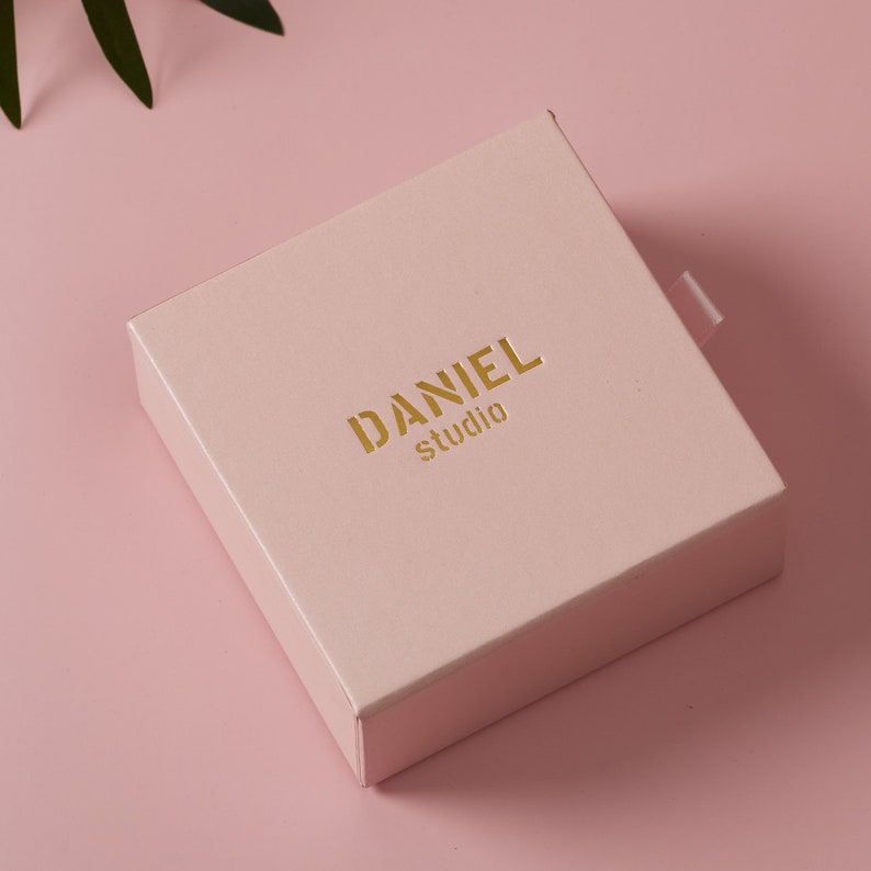 Może przedstawiać: Jasnor&oacute;żowe kwadratowe pudełko z złotym logo z napisem "DANIEL studio".