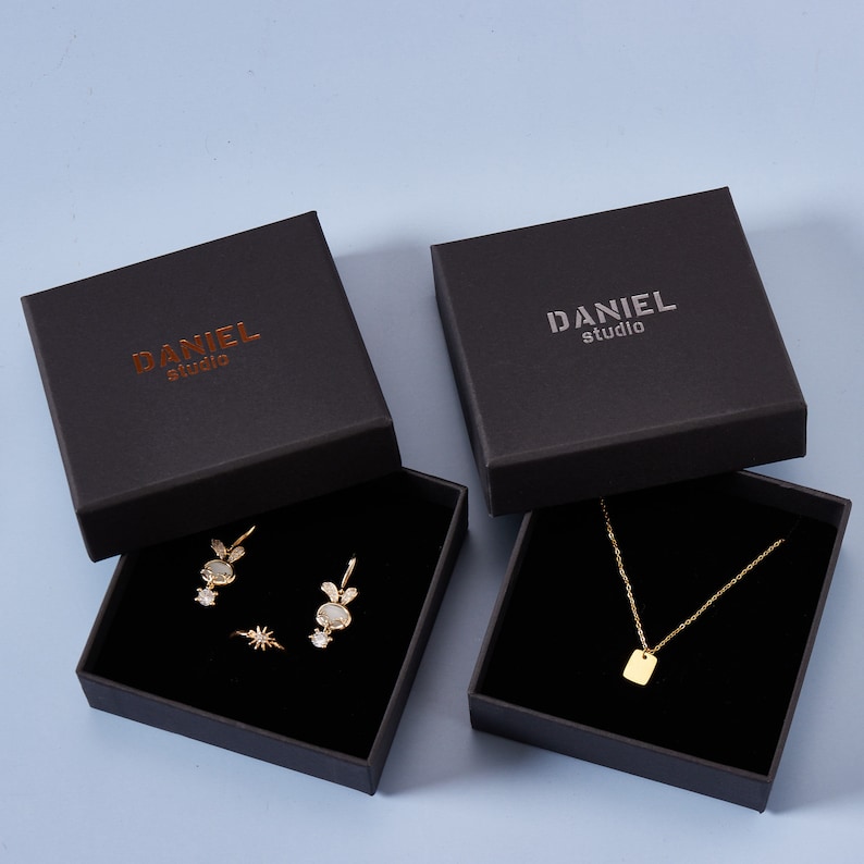 May include: Two black gift boxes with the text "DANIEL studio" on the top. One box contains a gold necklace with a square pendant. The other box contains a pair of gold earrings with a white stone and a gold ring with a star design.