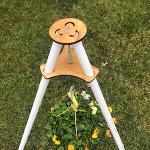 May include: A white hanging plant stand with three legs made of PVC pipe. The stand has a wooden top with a circular cutout and a screw for adjusting the height. A yellow hanging basket with yellow and white flowers is hanging from the stand.