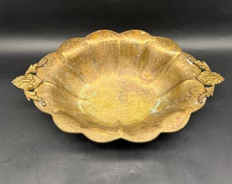 May include: A vintage brass bowl with a scalloped rim and ornate handles. The bowl has a hammered texture and a golden hue, with darker areas suggesting age and use. The handles feature detailed leaf-like designs.