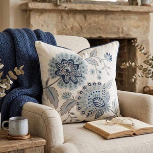 May include: A square throw pillow featuring an off-white fabric base with intricate embroidery in shades of navy blue, slate blue, and light gray. The design displays large stylized floral motifs with detailed petals and leaves, finished with a dark blue piped edge.
