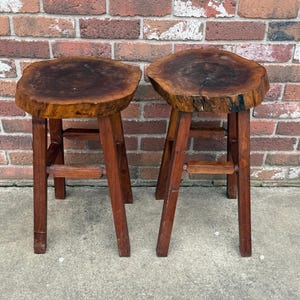 May include: Two wooden stools with a natural wood finish. The tops of the stools are made from a single piece of wood with a live edge. The stools have a simple design with three legs.
