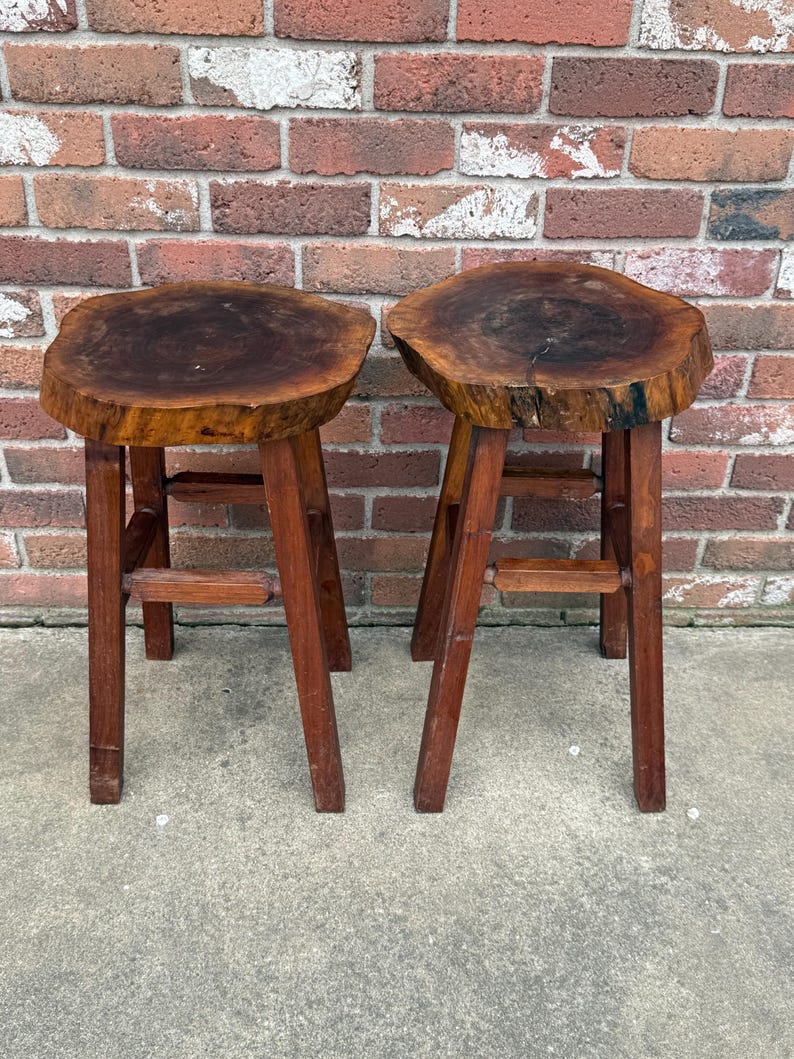 May include: Two wooden stools with a natural wood finish. The tops of the stools are made from a single piece of wood with a live edge. The stools have a simple design with three legs.