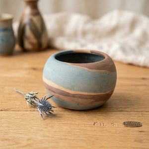 May include: A small, round, ceramic bowl featuring a marbled pattern of muted blue, tan, and brown tones. The bowl has a smooth, matte finish and a wide, open top. A dried, spiky blue thistle flower lies next to the bowl on a wooden surface.