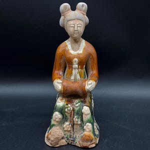 Könnte beinhalten: Eine Keramikfigur einer Frau in einem braunen und grünen Gewand, die eine Schriftrolle hält. Die Figur sitzt auf einem Sockel mit floralem Design.