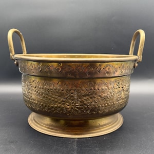 May include: A vintage brass planter with two handles and intricate floral designs. The planter has a wide base and a rounded body.