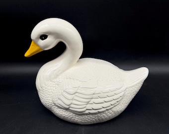 May include: A white ceramic swan figurine with a yellow beak and black eyes. The swan has detailed feather textures on its body and is set against a black background. This decorative piece is likely for home decor.