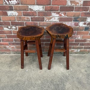 May include: Two wooden stools with a natural wood finish. The tops of the stools are made from a single piece of wood with a live edge. The stools have a simple design with four legs.