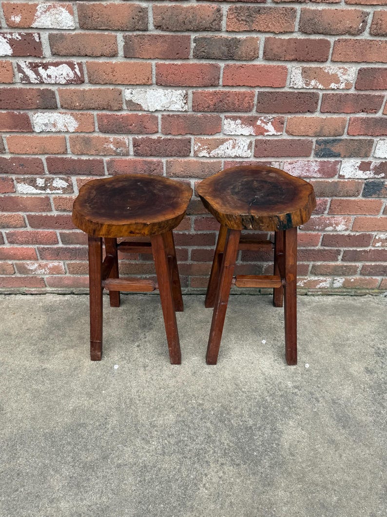 May include: Two wooden stools with a natural wood finish. The tops of the stools are made from a single piece of wood with a live edge. The stools have a simple design with four legs.