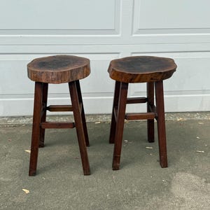 May include: Two wooden stools with a natural wood finish. The tops of the stools are made from a thick slice of tree trunk. The stools have a simple, rustic design.