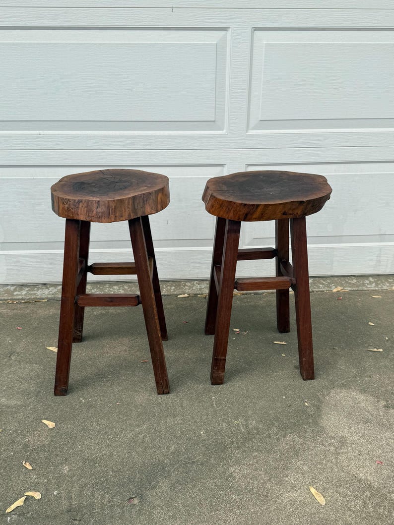 May include: Two wooden stools with a natural wood finish. The tops of the stools are made from a thick slice of tree trunk. The stools have a simple, rustic design.