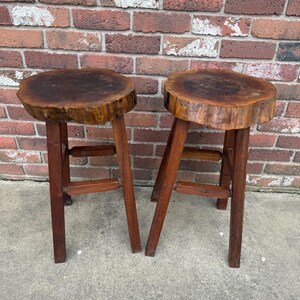 May include: Two wooden stools with a natural wood finish. The stools have a round, thick top that looks like a tree trunk slice. The stools have four legs and a simple, rustic design.
