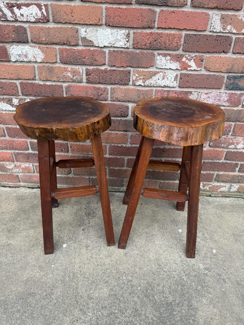 May include: Two wooden stools with a natural wood finish. The stools have a round, thick top that looks like a tree trunk slice. The stools have four legs and a simple, rustic design.