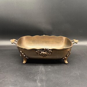 Puede incluir: Un cuenco decorativo rectangular de color bronce con un borde festoneado. El cuenco presenta asas ornamentadas y está sostenido por cuatro patas decorativas. Un diseño de racimo de uvas adorna la parte delantera. Probablemente vintage.