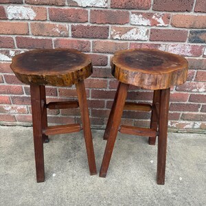 May include: Two wooden stools with round, natural wood tops and simple, sturdy legs. The tops are made from tree slices, showing the natural grain and texture of the wood.