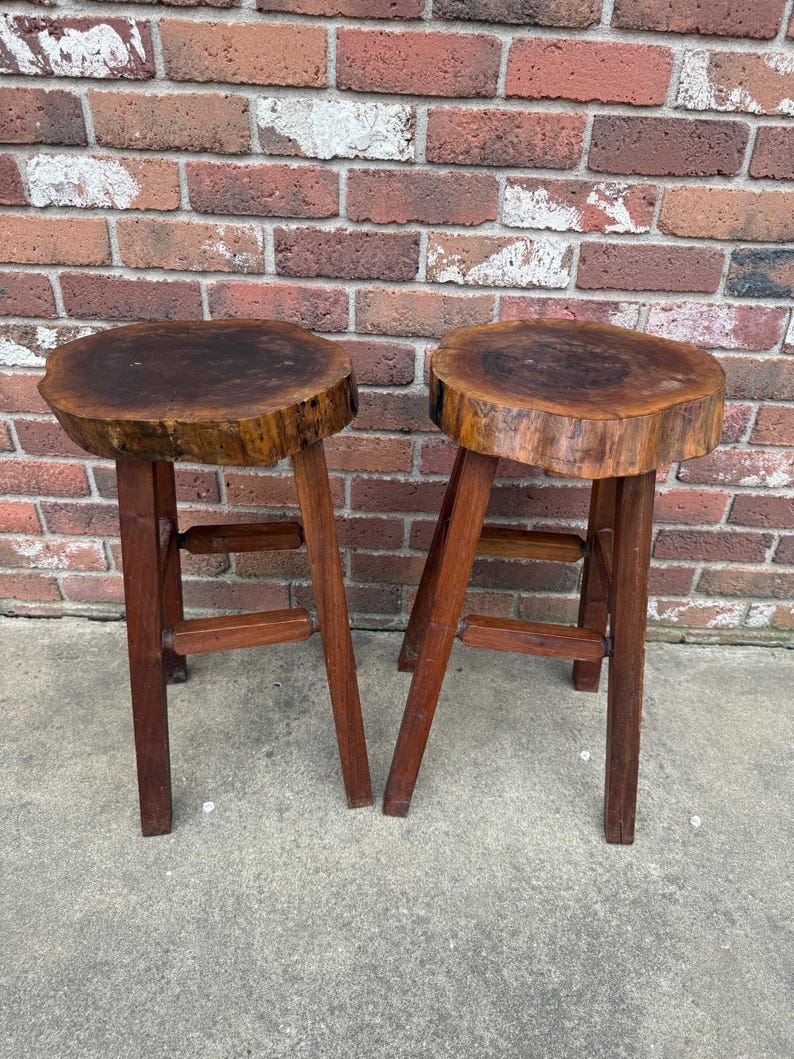 May include: Two wooden stools with round, natural wood tops and simple, sturdy legs. The tops are made from tree slices, showing the natural grain and texture of the wood.