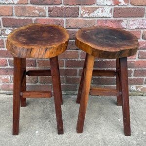 May include: Two wooden stools with a natural wood finish. The tops of the stools are made from a single piece of wood with a live edge. The stools have three legs each.