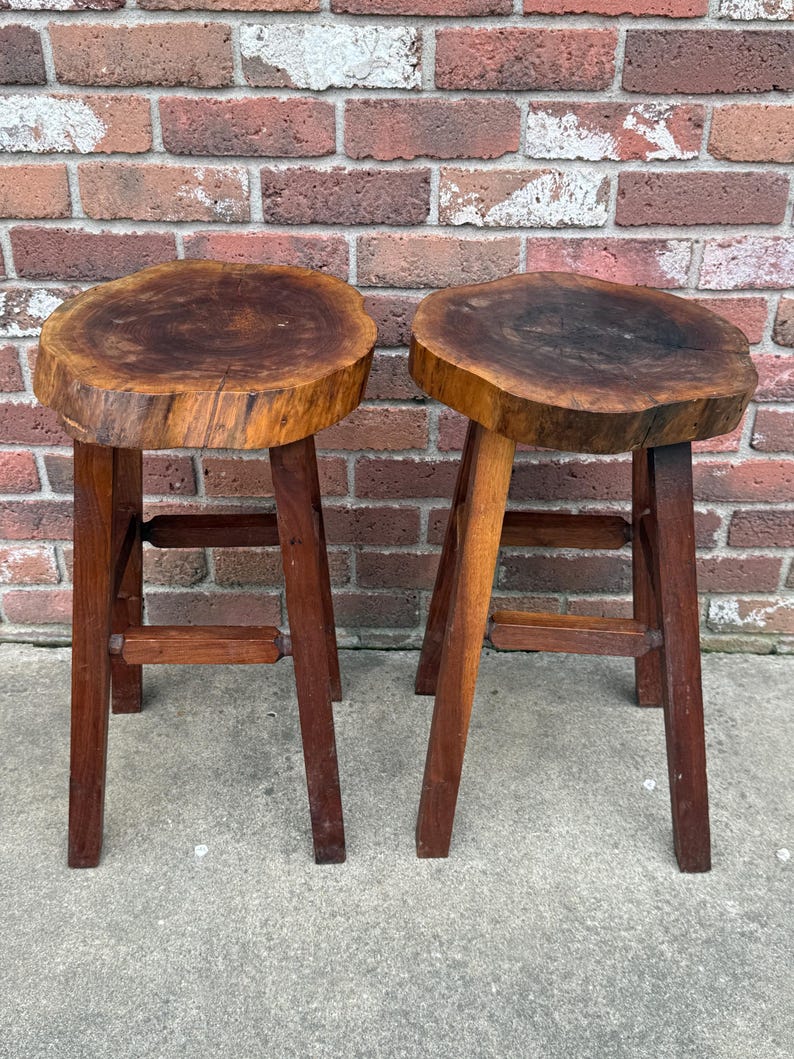May include: Two wooden stools with a natural wood finish. The tops of the stools are made from a single piece of wood with a live edge. The stools have three legs each.