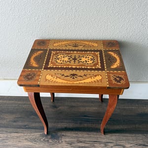 May include: A rectangular wooden side table featuring intricate marquetry inlay on the tabletop. The surface displays a central panel with floral motifs surrounded by geometric borders in shades of light, medium, and dark brown wood. It stands on four curved legs.