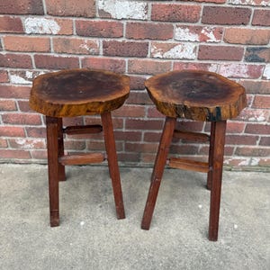 May include: Two wooden stools with round, natural wood tops and simple, sturdy legs. The stools are made from dark wood and have a rustic, handcrafted look.