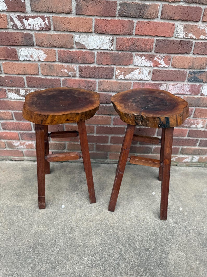 May include: Two wooden stools with round, natural wood tops and simple, sturdy legs. The stools are made from dark wood and have a rustic, handcrafted look.