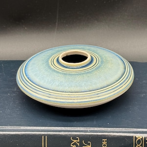 May include: A small, round, blue and white ceramic vase with a wide opening. The vase has a glazed finish and a subtle, textured pattern.