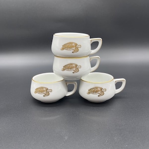 Könnte beinhalten: Set aus vier weißen Keramik-Teetassen mit goldenen Rändern und einem braunen Schildkröten-Design.